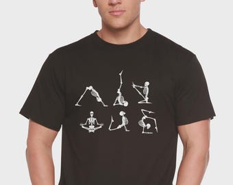 May include: Black t-shirt featuring a white graphic of skeletons in various yoga poses. The design includes poses like downward dog, handstand, and seated meditation. The shirt is made of a soft material.