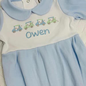 May include: Baby blue and white romper with a Peter Pan collar. The romper features embroidered golf carts in shades of green and blue, with the name "Owen" embroidered in blue thread.
