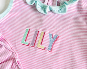May include: Pink and white striped top with the name "LILY" embroidered in colorful letters. The top has a light blue trim and a ruffled collar. The fabric appears to be soft and lightweight, suitable for warm weather.