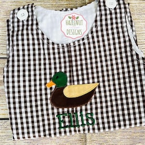 May include: Brown and white gingham baby romper with a duck applique. The duck has a green head, yellow wing, and orange beak. The name "Ellis" is embroidered in green below the duck. The romper has white buttons and a white lining.