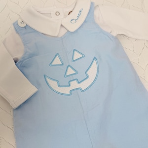 May include: A light blue baby outfit featuring a jack-o'-lantern face design. The outfit includes a long-sleeved white shirt and a blue corduroy overall with a smiling pumpkin face. The name "Owen" is embroidered on the collar.