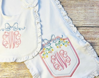 May include: A white baby bib and burp cloth set with ruffled edges. Both pieces feature embroidered floral designs and a monogrammed "EVE" in pink thread.