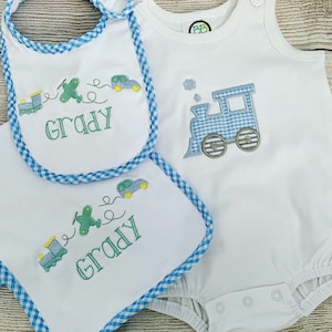 May include: A white baby bodysuit with a blue and white gingham train design. Two white bibs with blue gingham trim feature a green airplane, car, and train design with the name "Grady" embroidered on each.