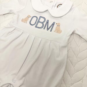 May include: White baby romper with a Peter Pan collar and long sleeves. The romper features embroidered tan-colored dog illustrations flanking the blue embroidered letters "OBM". The garment has pleats at the waist and snap closures.