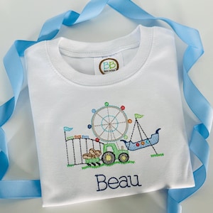 May include: White t-shirt with an embroidered carnival scene, including a Ferris wheel, swing ride, and tractor. The name "Beau" is embroidered in navy blue script below the design. A light blue ribbon is draped around the shirt.