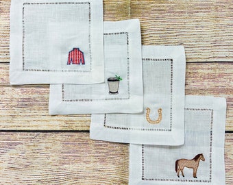 May include: Four white linen napkins with embroidered designs. The designs include a red and white striped shirt, a cocktail glass with a green garnish, a horseshoe, and a brown horse.