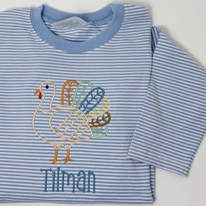 May include: A long-sleeve shirt with light blue and white horizontal stripes. The shirt features an embroidered turkey with colorful feathers in orange, teal, and blue, and the name Tilman embroidered in blue thread below the turkey.