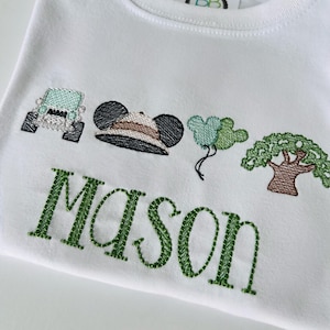 May include: White t-shirt with green embroidered text "Mason" and various embroidered images: a light blue jeep, a safari hat, green balloons, and a tree. The shirt has a round neckline.