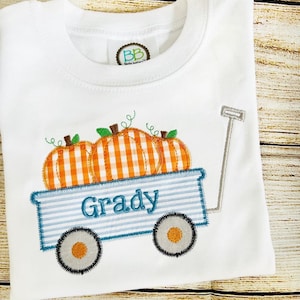 May include: White baby onesie with an embroidered design of a blue wagon with three orange and white gingham pumpkins and the name "Grady" on the side.