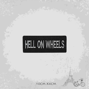 May include: Black embroidered patch with white text that reads "Hell on Wheels". The patch is 10 cm by 4 cm.