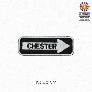 Op de afbeelding: Zwart-witte geborduurde strijkpatch met een witte pijl die naar rechts wijst en het woord "CHESTER" in witte tekst. De patch is 7,5 cm bij 3 cm.