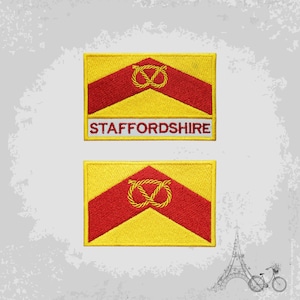 May include: Two embroidered patches featuring the flag of Staffordshire, England. The flag is yellow with a red chevron and a red knot in the centre. The text "STAFFORDSHIRE" is written in red on a white background below the flag.
