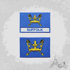 Könnte beinhalten: Zwei gestickte Aufnäher mit dem Wappen von Suffolk, England. Die Aufnäher sind blau mit einer goldenen Krone und zwei gekreuzten Pfeilen. Der obere Aufnäher trägt das Wort "SUFFOLK" in weiß unter dem Wappen.