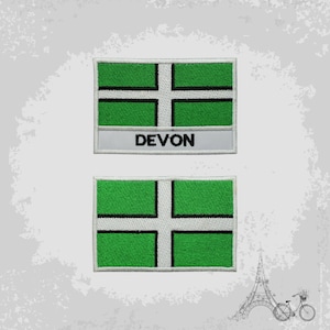May include: Two embroidered patches featuring the flag of Devon, England. The design includes a green field with a white cross and black border. One patch has the word "DEVON" embroidered in black on a white banner.