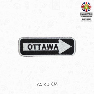 Puede incluir: Parche de hierro bordado en blanco y negro con una flecha blanca apuntando hacia la derecha y la palabra "OTTAWA" en texto blanco. El parche mide 7,5 cm por 3 cm.
