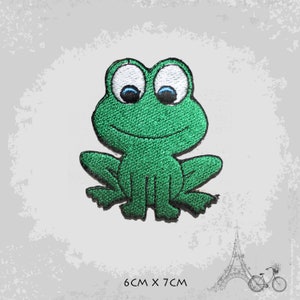 Może przedstawiać: Zielona haftowana łatka z żabą z białymi oczami i niebieskimi źrenicami. Łatka ma 6 cm na 7 cm.