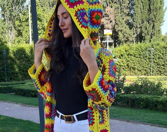 Puede incluir: Una larga chaqueta de ganchillo de colores con capucha. La chaqueta está hecha de cuadrados de la abuela en tonos de amarillo, rojo, azul, verde y morado. La modelo lleva pantalones blancos y zapatos negros.