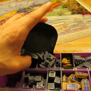 Könnte beinhalten: Eine violette Plastik-Aufbewahrungsbox mit mehreren Fächern, die mit verschiedenen Spielsteinen gefüllt sind. Die Fächer enthalten kleine graue Plastikteile, Holzteile und blaue und weiße Fensterteile.