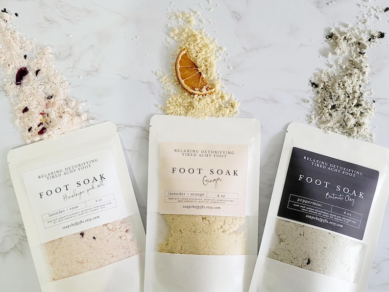 May include: Three white pouches with labels that read "Relaxing Detoxifying Tired Achy Foot Foot Soak" with different scents and ingredients. The scents are Himalayan pink salt, ginger, and bentonite clay. The pouches are filled with a white, beige, and gray powder.