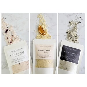 May include: Three white pouches with black text on the front, each containing a different foot soak. The pouches are labeled "Foot Soak" with the scents "Lavender + Orange", "Ginger", and "Peppermint + Charcoal".
