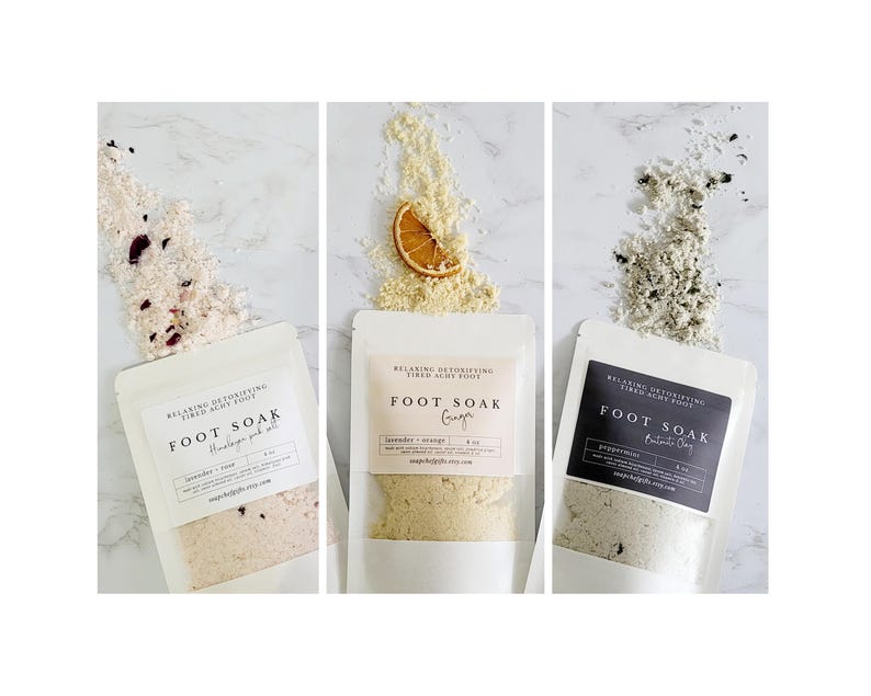 May include: Three white pouches with black text on the front, each containing a different foot soak. The pouches are labeled "Foot Soak" with the scents "Lavender + Orange", "Ginger", and "Peppermint + Charcoal".