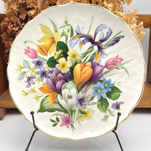 May include: A porcelain plate with a scalloped edge and gold trim, decorated with a colourful floral design. The arrangement includes daffodils, irises, tulips, and crocuses in yellow, purple, pink, and blue hues. The plate is displayed on a black metal stand.
