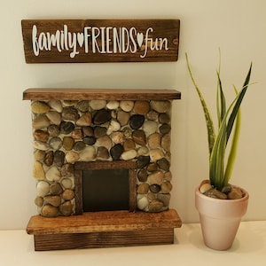 May include: A miniature stone fireplace with a wooden mantel and hearth. The fireplace has a sign above it that reads "family♥friends♥fun". A potted plant sits in front of the fireplace.