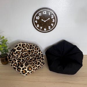May include: Two beanbag chairs, one with a leopard print and one black, are on a wooden surface. A brown wooden clock with white numbers and hands is on the wall behind the chairs.