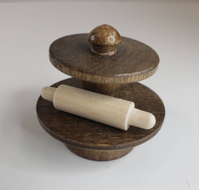 Puede incluir: Un rodillo de madera y un soporte de madera con una parte superior redonda. El soporte tiene un peque&ntilde;o pomo en la parte superior. El rodillo est&aacute; apoyado en el soporte.