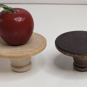 May include: Two wooden pedestal stands, one with a red apple on top. The stand on the left is light wood with a natural finish. The stand on the right is dark brown wood with a distressed finish.