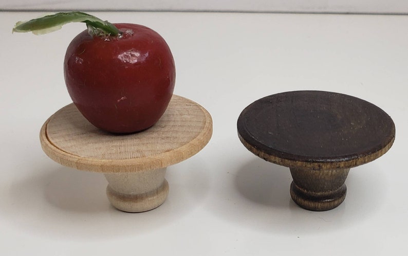 May include: Two wooden pedestal stands, one with a red apple on top. The stand on the left is light wood with a natural finish. The stand on the right is dark brown wood with a distressed finish.