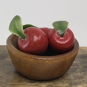 May include: A small wooden bowl filled with three red apples with green leaves. The bowl is on a wooden surface.