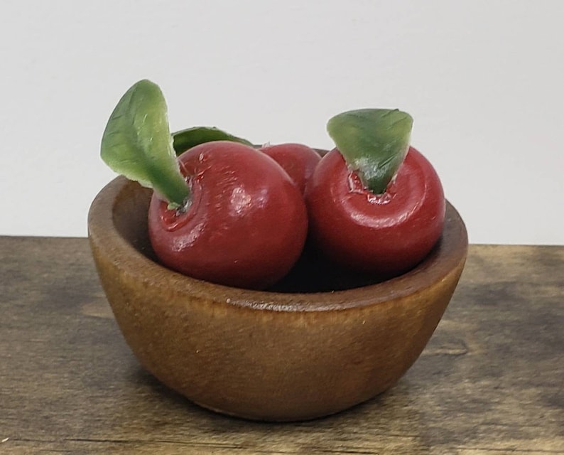 May include: A small wooden bowl filled with three red apples with green leaves. The bowl is on a wooden surface.