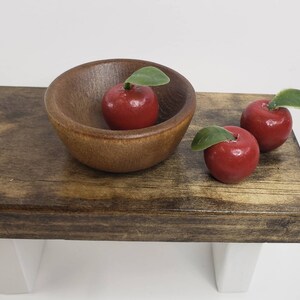 May include: Three red apples with green leaves sit on a wooden table. One apple is in a brown wooden bowl.