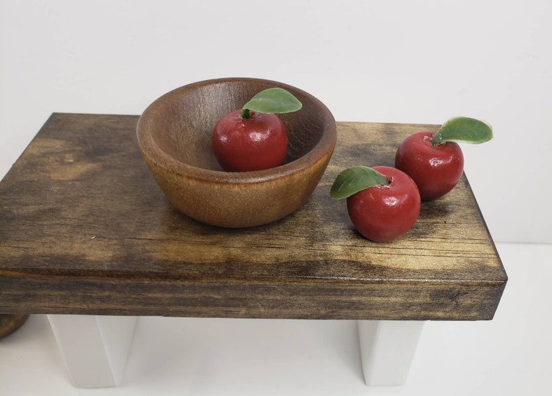 May include: Three red apples with green leaves sit on a wooden table. One apple is in a brown wooden bowl.