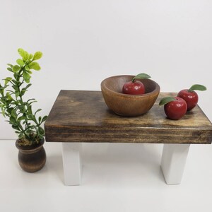 May include: A wooden table with white legs and a brown wooden top. There is a wooden bowl with three red apples on the table.