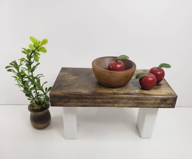 May include: A wooden table with white legs and a brown wooden top. There is a wooden bowl with three red apples on the table.
