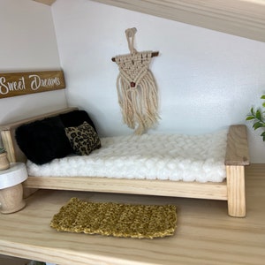 May include: A dollhouse bedroom with a white bed, a wooden headboard, and a white fluffy blanket. There are two pillows on the bed, one black and one with a leopard print. A small wooden side table with a white top sits next to the bed. A small potted plant sits on the table. A wooden sign with the words "Sweet Dreams" is on the wall above the bed. A macrame wall hanging with a red bead hangs on the wall above the bed. A small rug with a gold color sits on the floor in front of the bed.