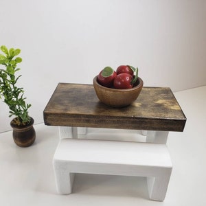 May include: A miniature wooden table and bench set with a white bench and a brown table top. A wooden bowl filled with red apples sits on the table.