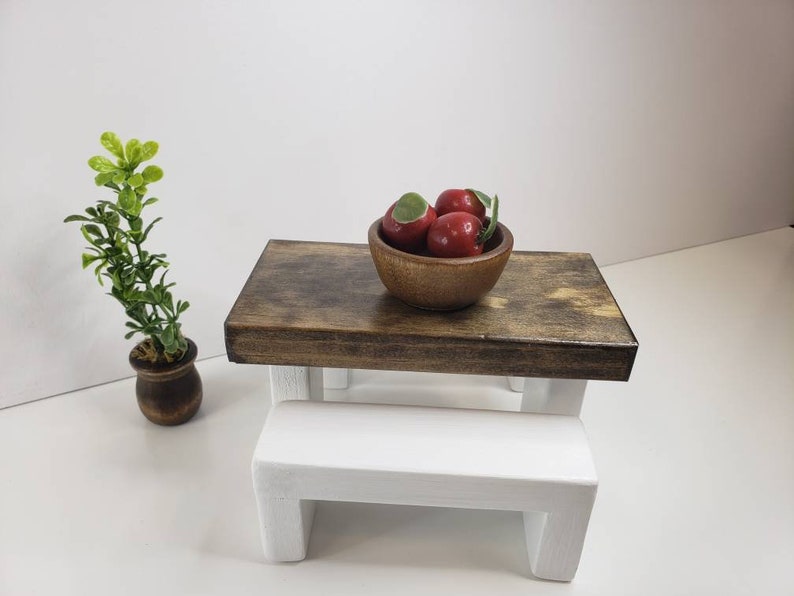 May include: A miniature wooden table and bench set with a white bench and a brown table top. A wooden bowl filled with red apples sits on the table.