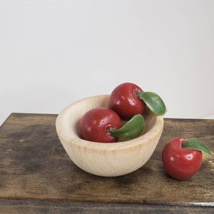 May include: A small wooden bowl filled with three red apples with green leaves. The bowl is sitting on a wooden surface.