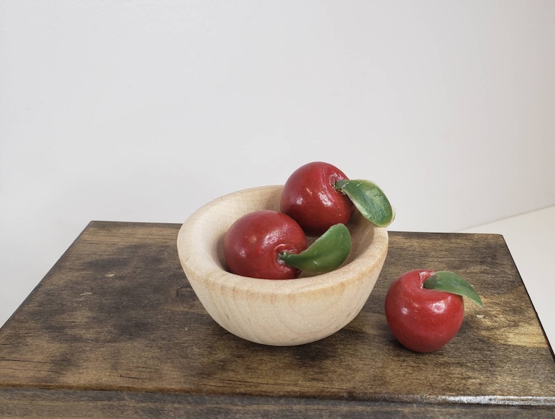May include: A small wooden bowl filled with three red apples with green leaves. The bowl is sitting on a wooden surface.