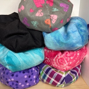May include: A stack of seven fabric-covered cushions in various colors and patterns, including gray with hearts, blue with swirls, turquoise with swirls, pink with a floral pattern, purple with white polka dots, and purple and green plaid.