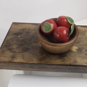 May include: A wooden table top with a small wooden bowl filled with three red apples. The bowl is in the center of the table top.