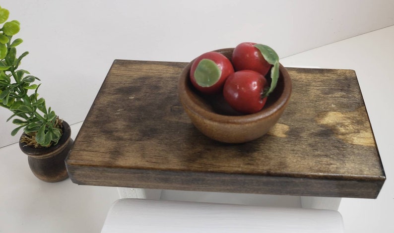 May include: A wooden table top with a small wooden bowl filled with three red apples. The bowl is in the center of the table top.