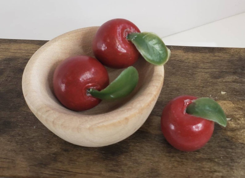 May include: Three red apples with green leaves in a small wooden bowl. The bowl is sitting on a wooden surface.