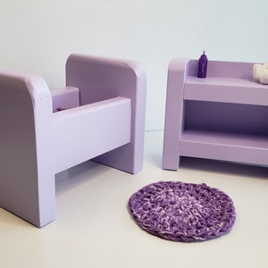 May include: A lavender wooden doll crib, a lavender wooden doll changing table with a shelf, and a lavender crocheted rug. The changing table has a small bottle and a white teddy bear on top.