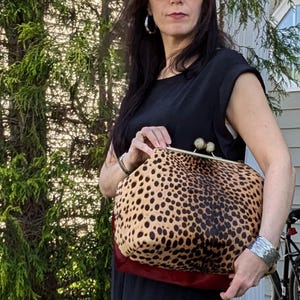 May include: A leopard print clutch bag with a gold-toned clasp. The bag has a tan and black spotted pattern, a red base, and a gold frame. The woman in the image is wearing sunglasses and a black dress.