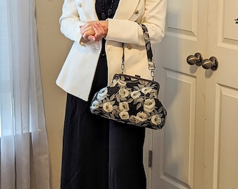 May include: A woman wearing a cream-colored blazer with gold buttons, a black top, and black wide-leg pants. She is carrying a black handbag with a floral pattern. The handbag has a black strap and a metal clasp. She is wearing black strappy sandals.