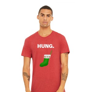 May include: A red heather t-shirt with the word "HUNG." printed in white. A green Christmas stocking with a white cuff and brown trim is hanging below the text.